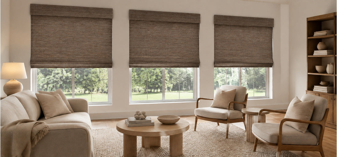 A beautifully styled, warm-toned living room features a beige sofa and wooden armchairs arranged around a round coffee table, with three large windows partially covered by textured brown woven shades.